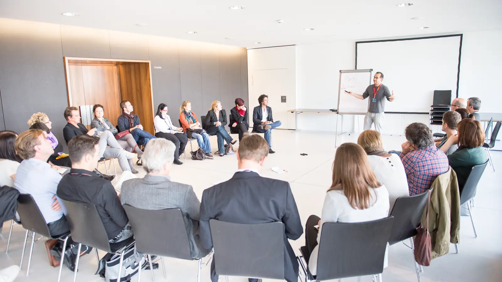 © Matthias Rhomberg Workshopteilnehmer:innen in einem Stuhlkreis und einem Vortragenden mit Flipchart und Leinwand in der weißen Seegalerie © Matthias Rhomberg