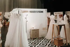 © The CLICK. Wedding Mehrere Brautkleider hängen und stehen auf einer Ausstellungsfläche mit Teppich und Möbeln. © The CLICK. Wedding