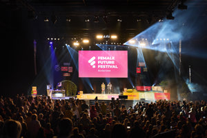 Vollbesetzte Reihenbestuhlung, eine Bühne mit Lounge-Möbeln und Leinwand in der Werkstattbühne des Festspielhauses Bregenz